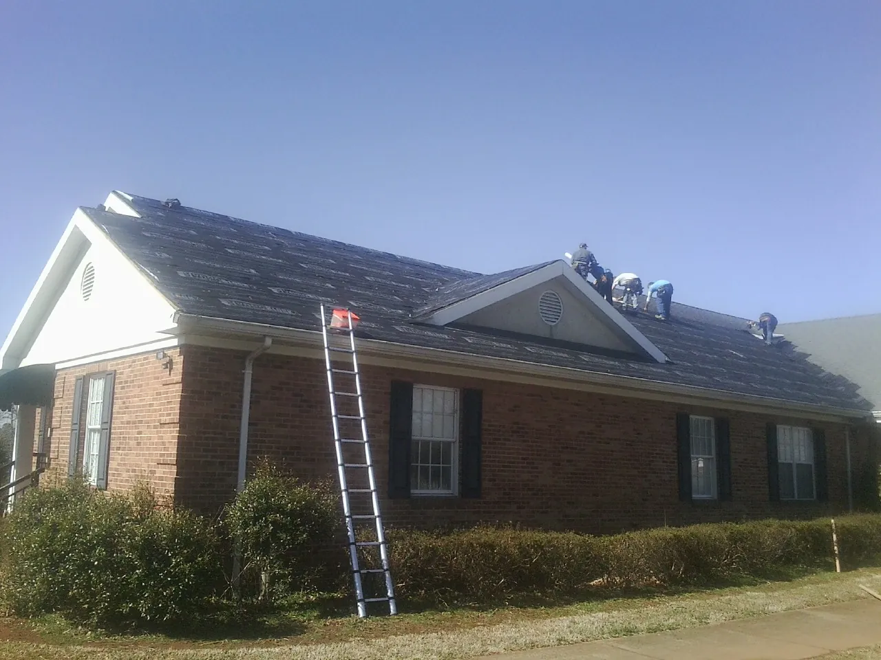 Crew tearing shingles off a brick ranch roof with a ladder set — Emergency Roof Repair work by our crew in Forsyth