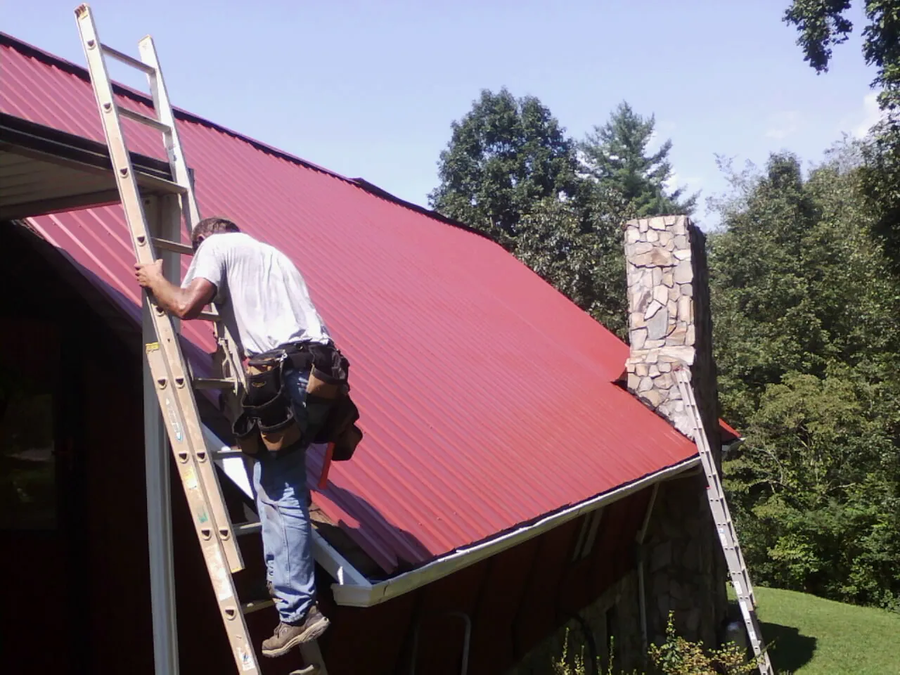 Roofer on a ladder beside a red metal roof and stone chimney — Soffit & Fascia Repair by Roofing Brothers in Forsyth